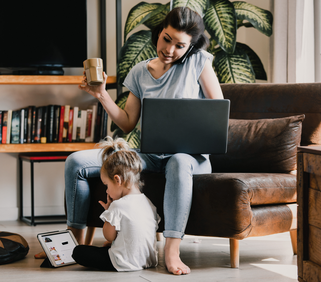 Mutter arbeitet mit Laptop auf dem Sofa, während ihr Kind auf dem Boden spielt